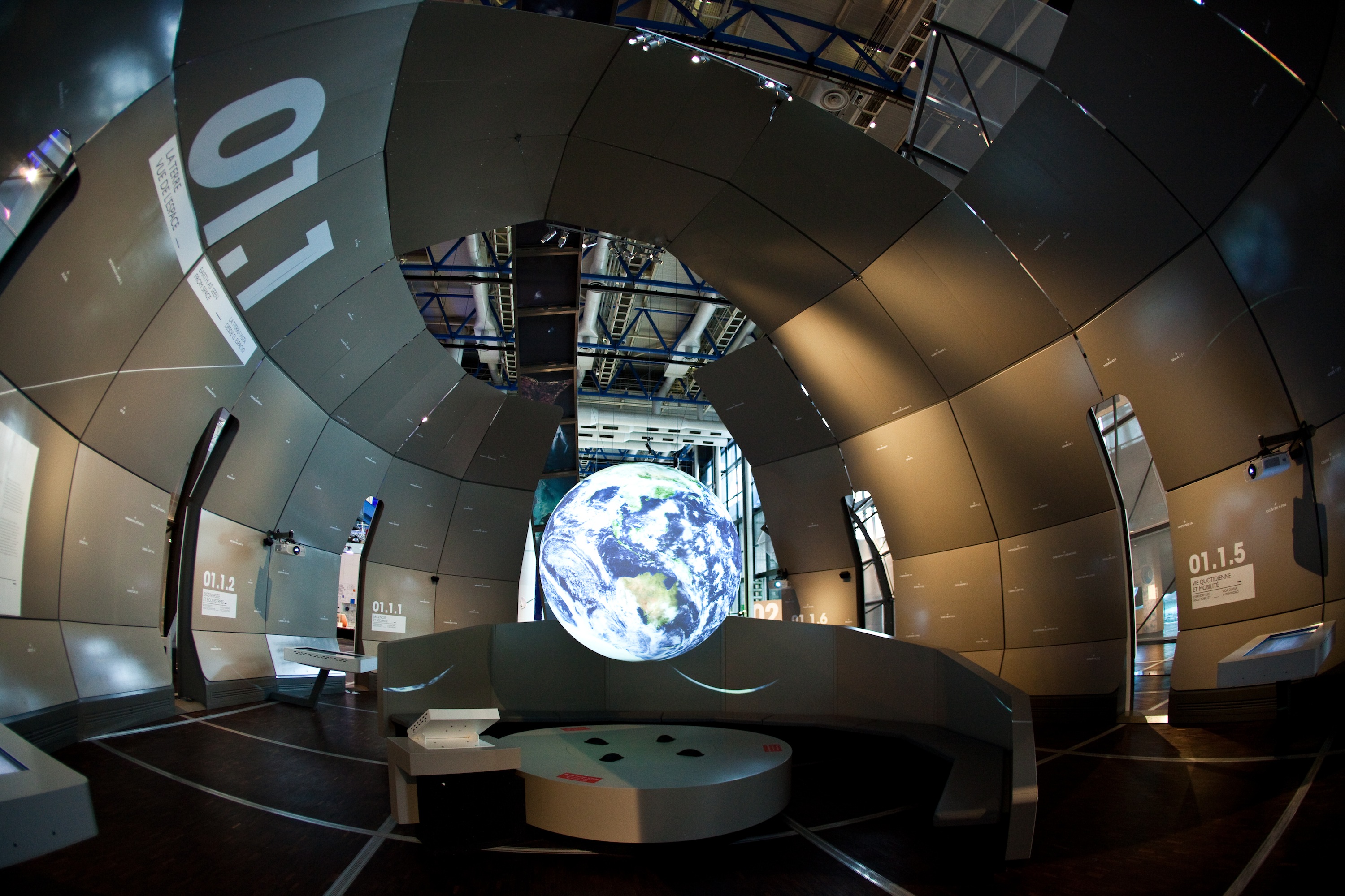 Science On a Sphere hangs from a domed ceiling constructed of individual panels displaying text. The dome is not fully enclosed, and through the gaps can be seen the ceiling of a larger room